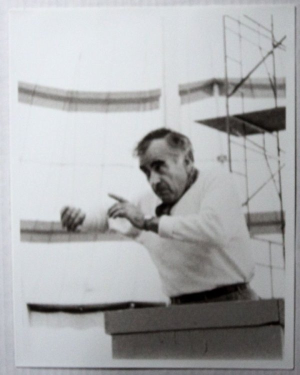 A LIVELY BLACK & WHITE PORTRAIT OF THE MEXICAN COMPOSER & CONDUCTOR CARLOS CHAVEZ REHEARSING ON AN OUTDOOR STAGE IN FRONT OF SCAFFOLDING.